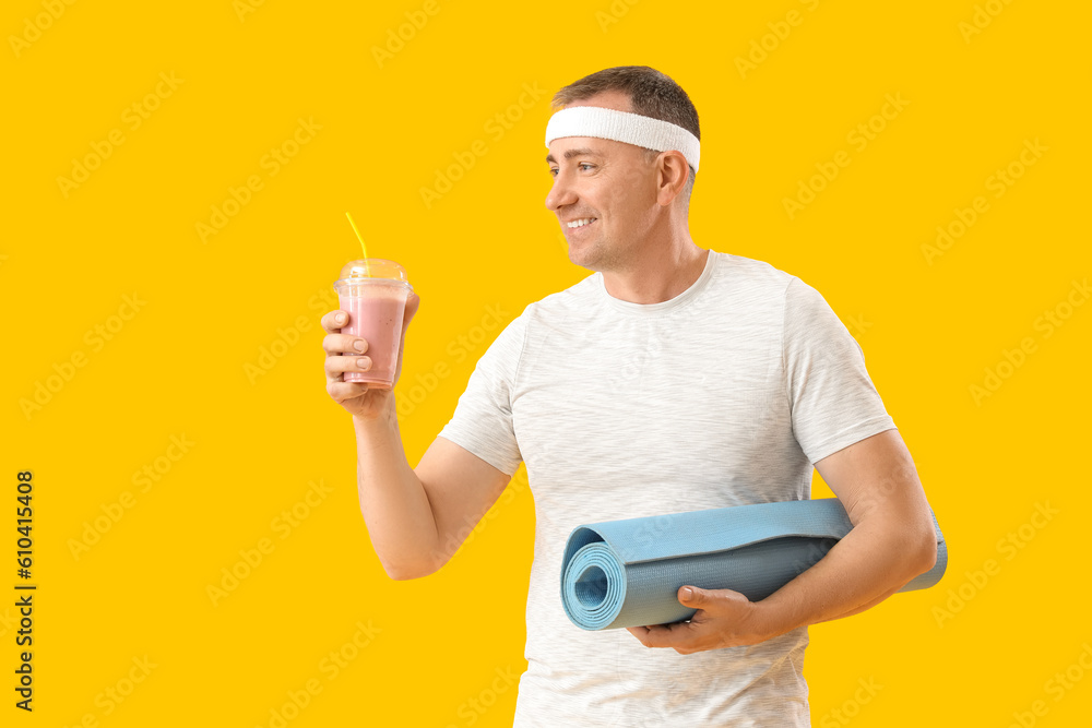 Sporty mature man with glass of fruit smoothie and fitness mat on yellow background