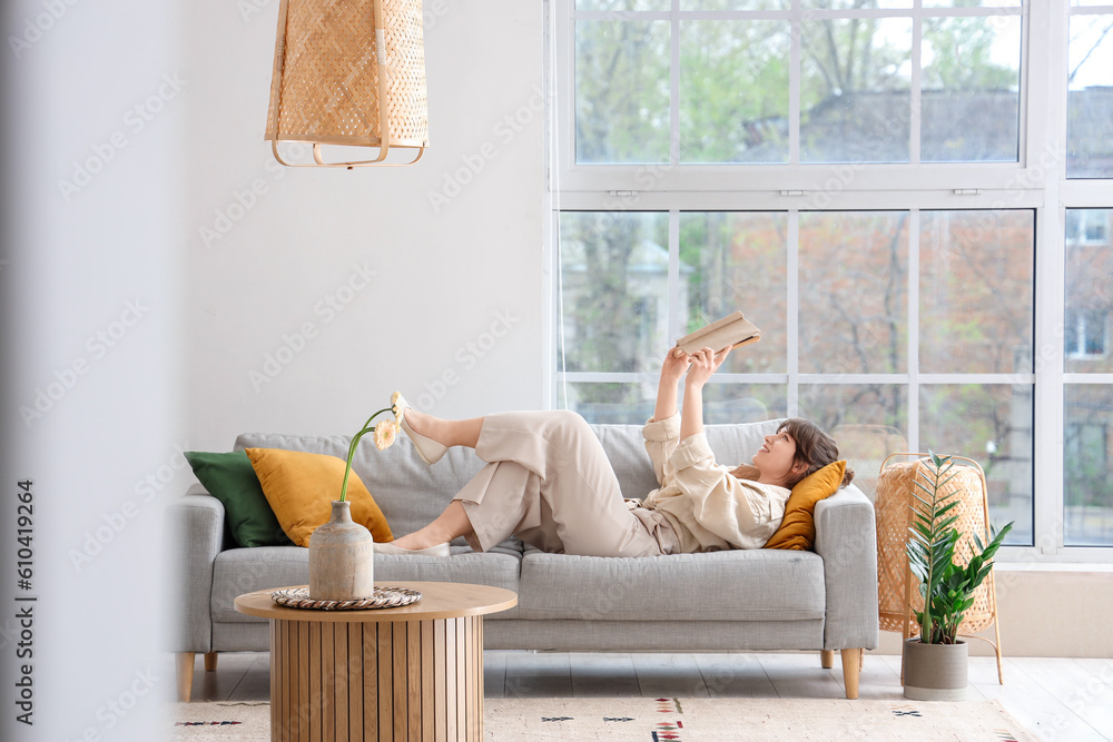 Happy young woman lying on grey sofa and reading book in interior of light living room, view from ha