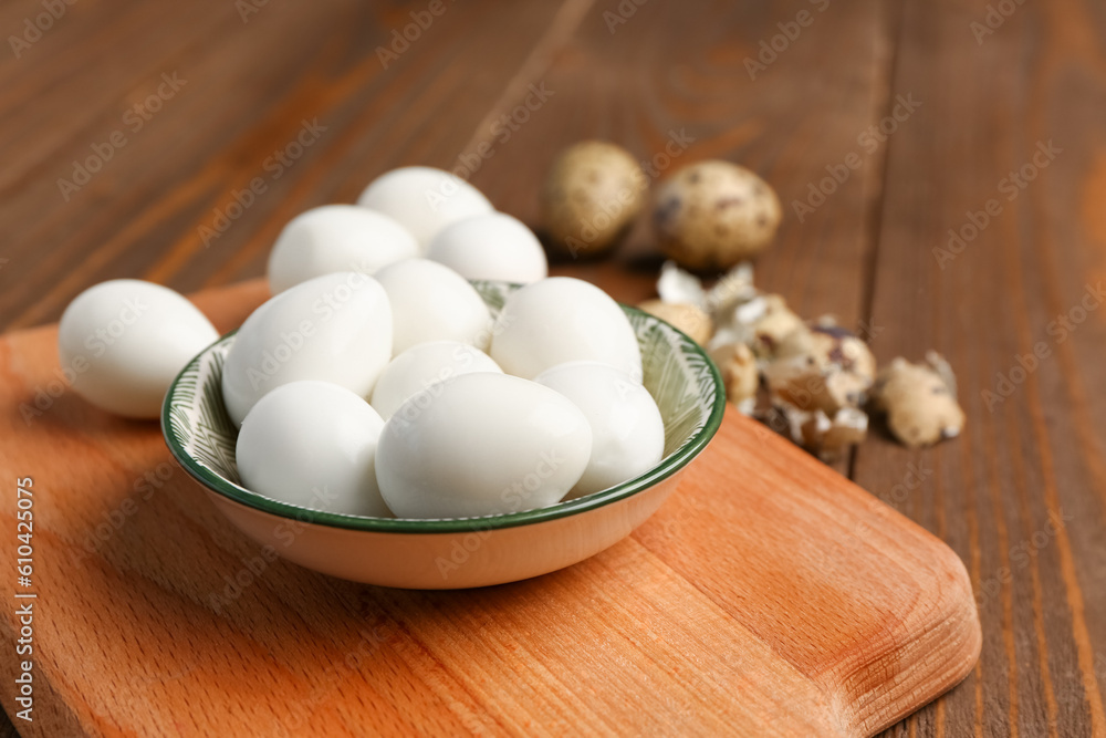 Board with bowl of boiled quail eggs and shells on wooden background