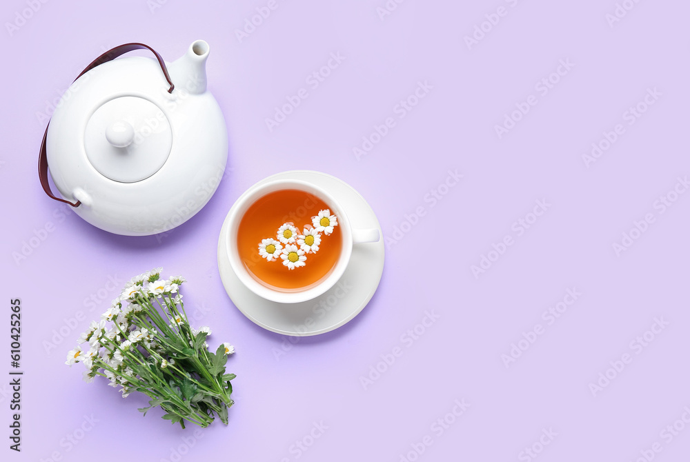 Teapot with cup of natural chamomile tea and flowers on lilac background