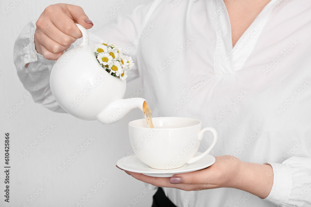 Beautiful young woman pouring chamomile tea from teapot into cup near white wall