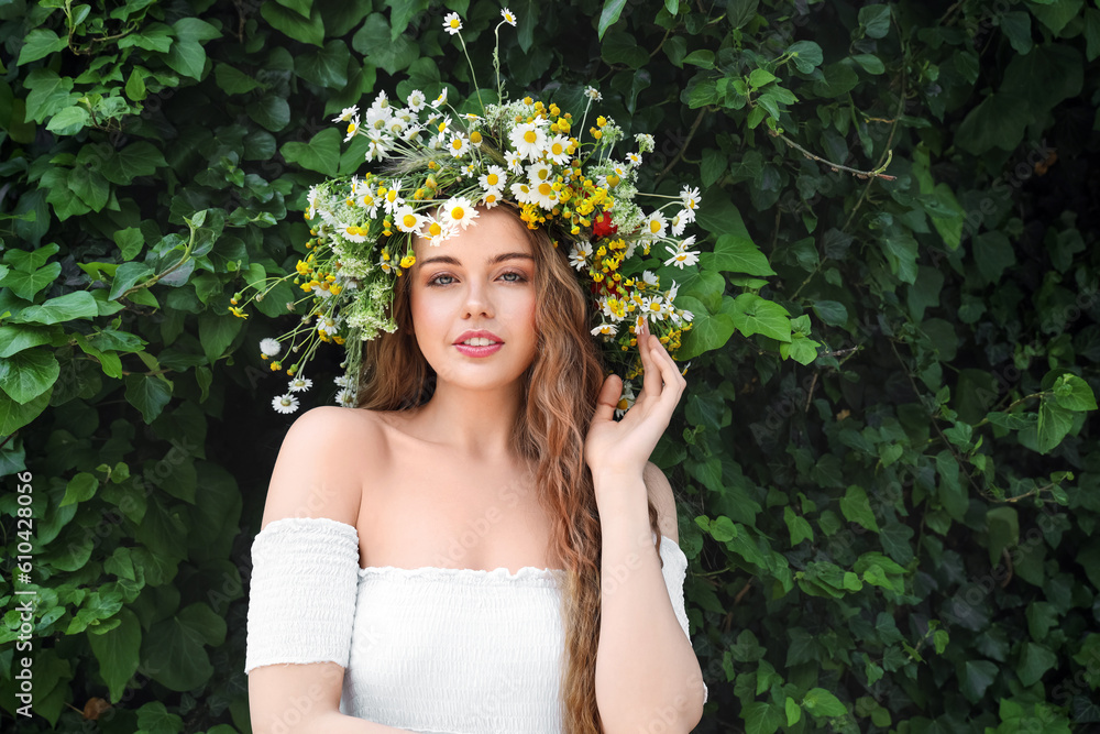 Beautiful young woman in flower wreath near green ivy outdoors. Summer solstice