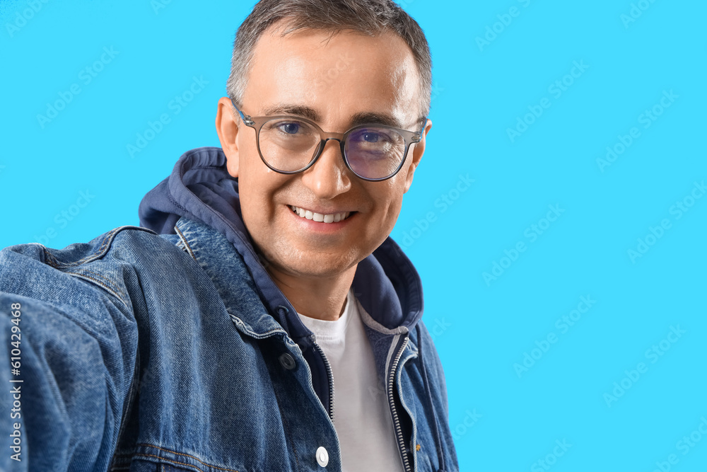 Mature man in eyeglasses taking selfie on blue background, closeup