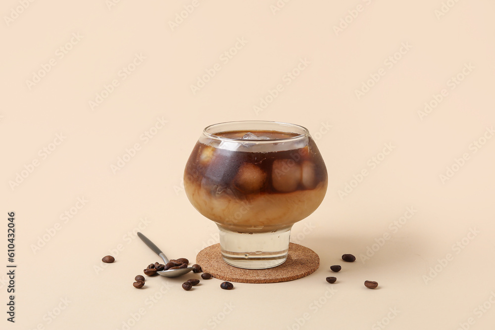 Glass of ice coffee with beans and spoon on beige background