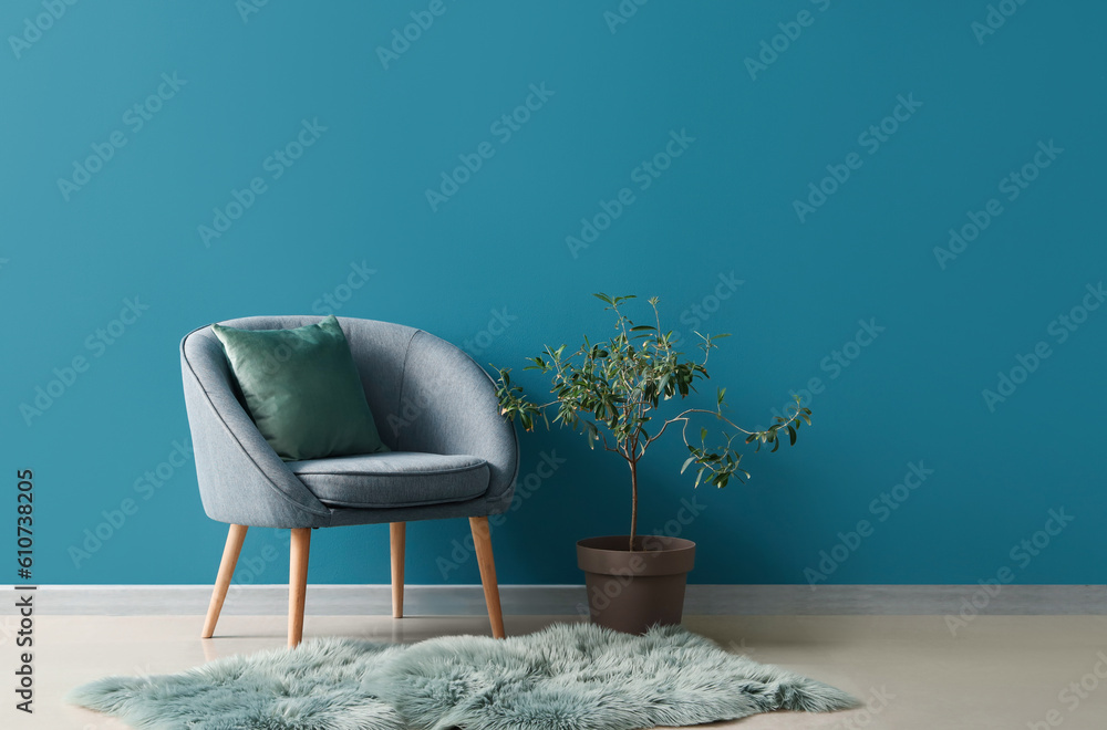 Cozy armchair with cushion, fur rug and houseplant on stool near blue wall