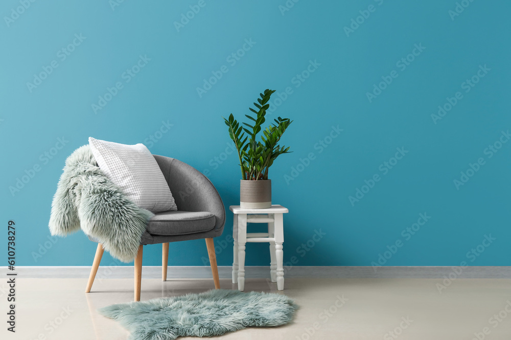 Cozy armchair with cushion, fur rug and houseplant on stool near blue wall