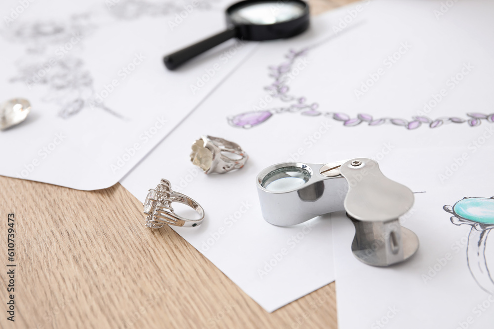 Rings with jewelers magnifier and drawings on wooden table, closeup