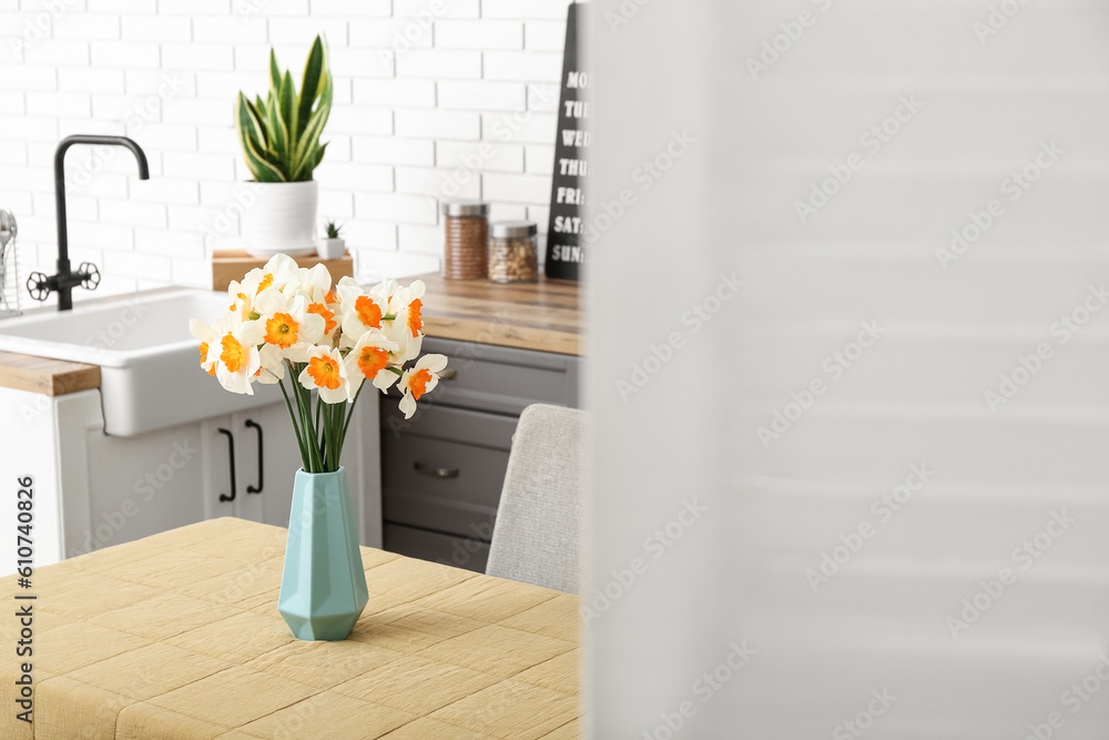 Vase with blooming narcissus flowers on table in modern kitchen, view behind folding screen