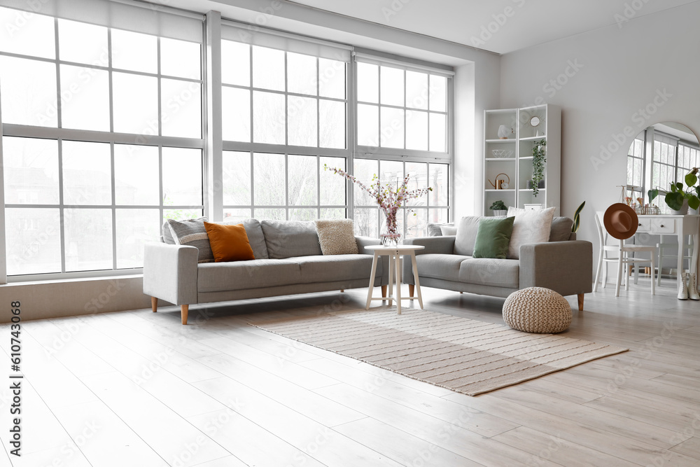 Interior of living room with grey sofas and blooming sakura branches on coffee table near big window