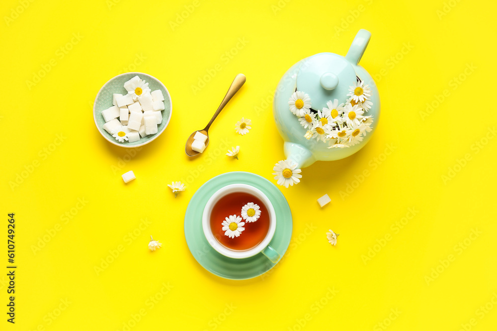 Teapot with cup of natural chamomile tea, sugar cubes and flowers on yellow background