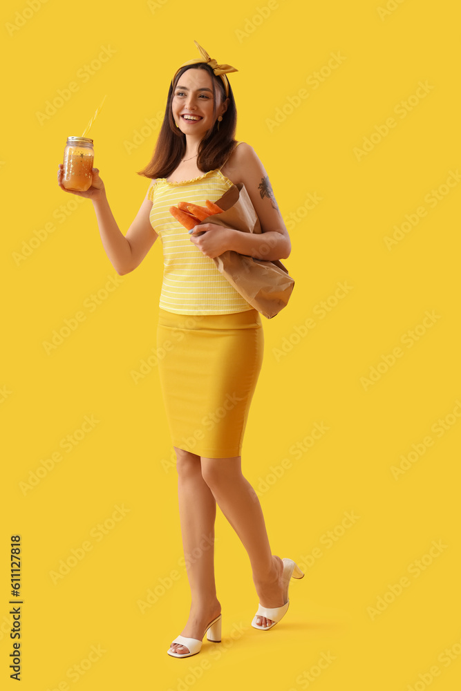 Young woman with glass jar of vegetable juice and carrots on yellow background