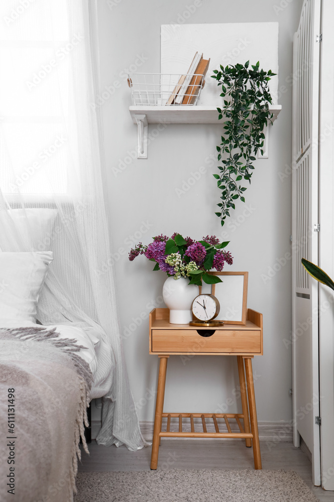 Vase with lilac flowers, clock and frame on table in bedroom