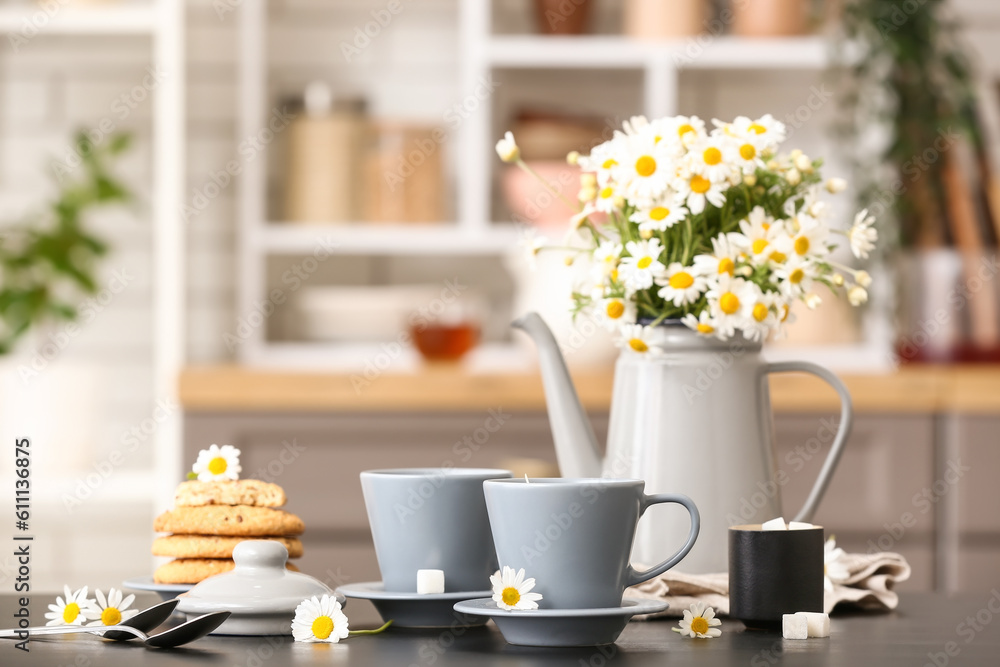 Watering can with flowers, cups of natural chamomile tea and cookies on black table in kitchen