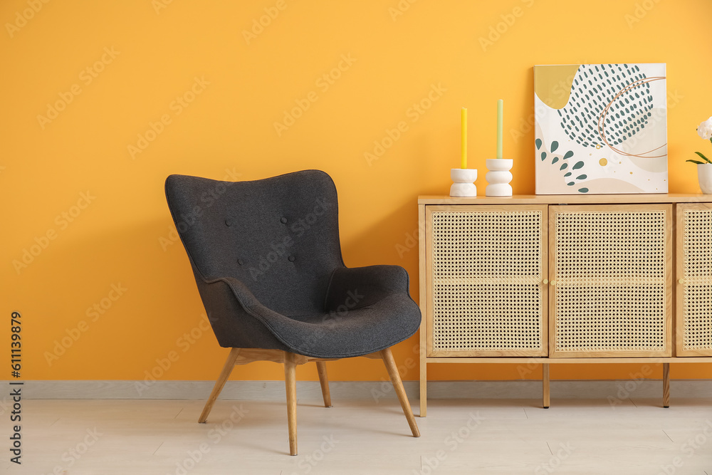 Dark grey armchair with dresser, painting and candles near orange wall