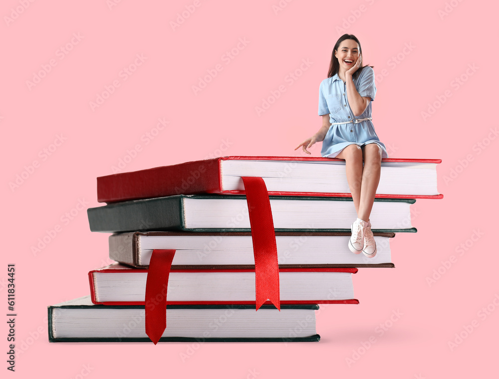 Happy young woman sitting on stack of big books against pink background