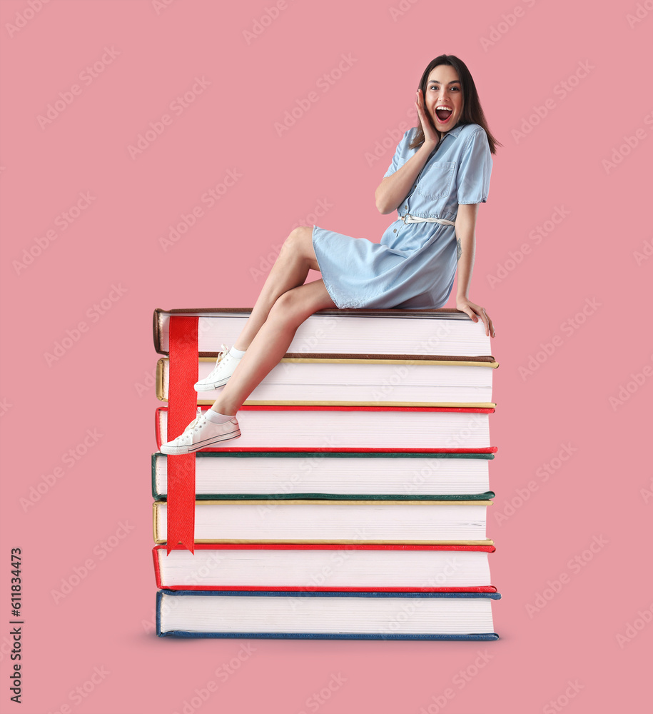 Happy young woman sitting on stack of big books against pink background