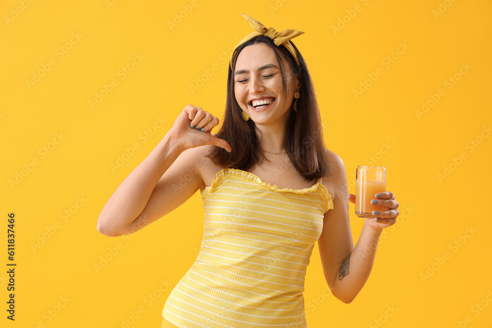 Young woman with glass of vegetable juice pointing at herself on yellow background