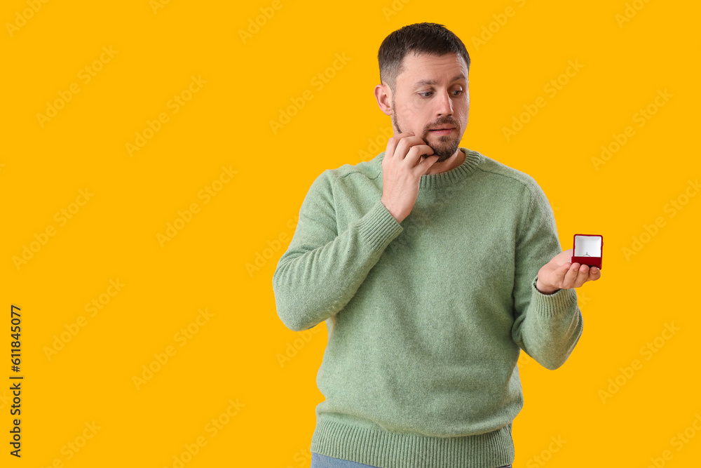 Worried young man with engagement ring on yellow background