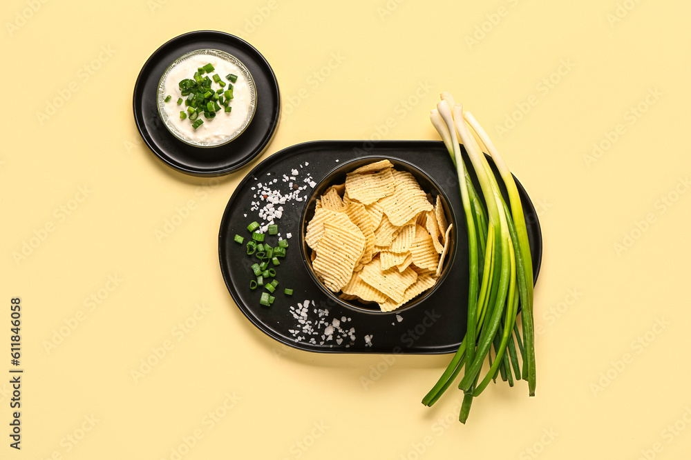 Bowl of tasty sour cream with sliced green onion and potato chips on yellow background