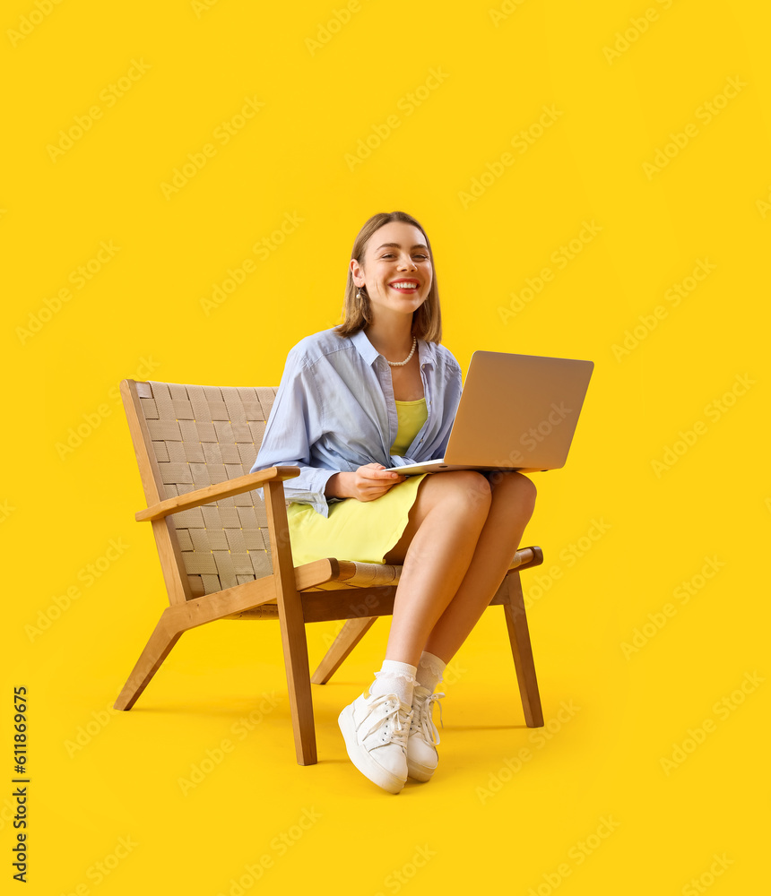 Young woman with laptop sitting in wooden armchair on yellow background