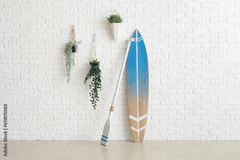 Wooden surfboard with paddle and houseplants on white brick wall in room