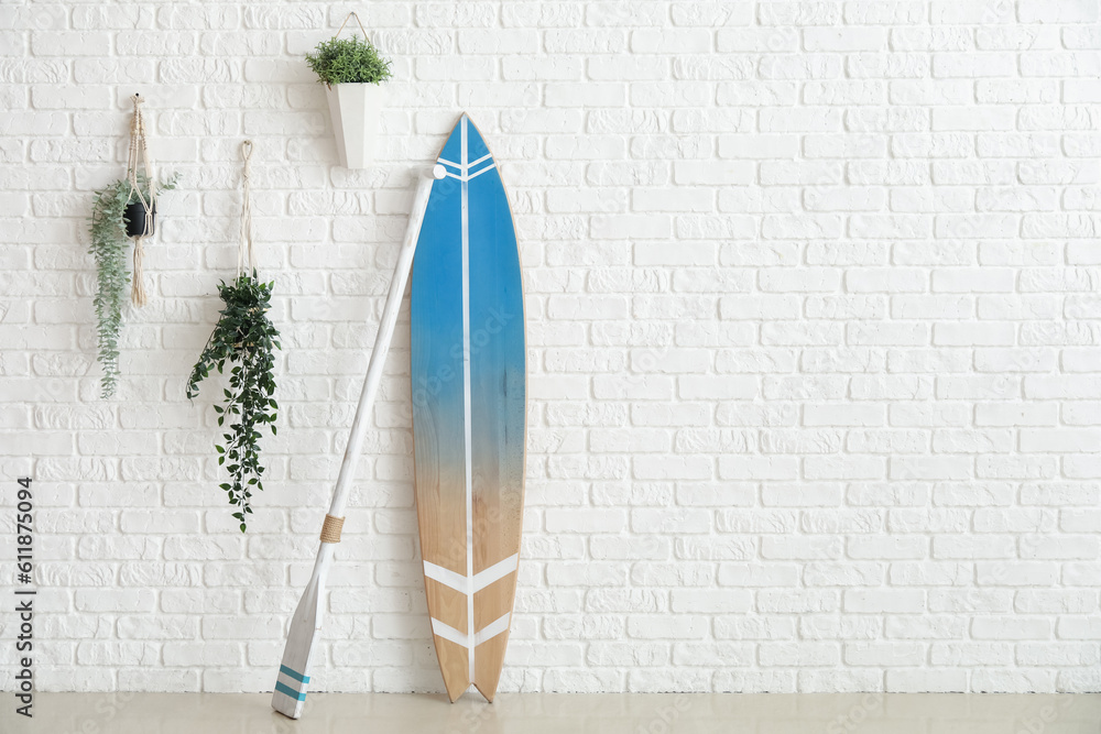 Wooden surfboard with paddle and houseplants on white brick wall in room