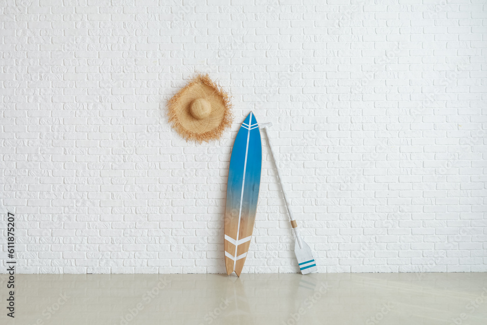 Wooden surfboard with paddle and straw hat on white brick wall in room