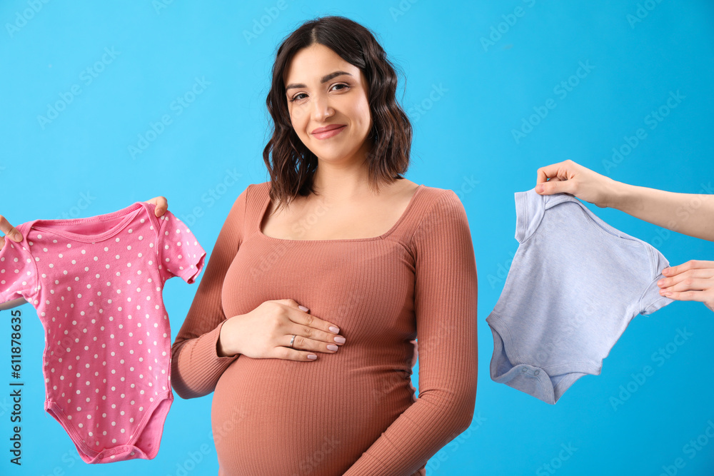 Young pregnant woman and female hands with baby bodysuits on blue background