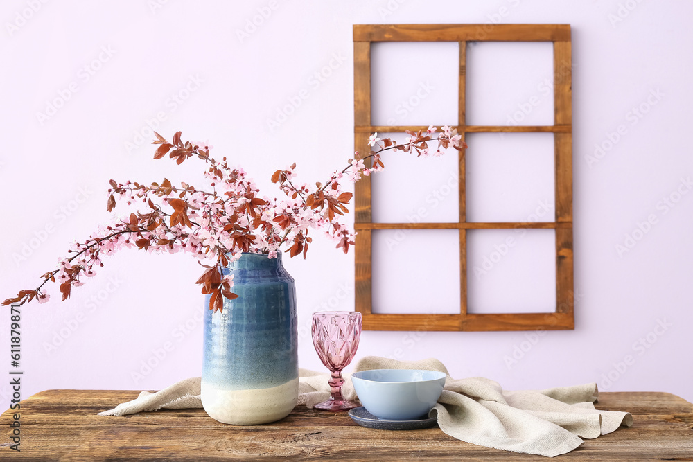 Vase with blooming tree branches, glass, bowl and napkin on wooden table near pink wall