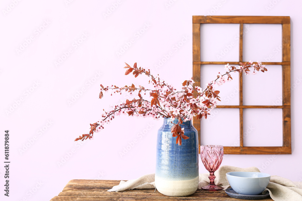 Vase with blooming tree branches, glass, bowl and napkin on wooden table near pink wall