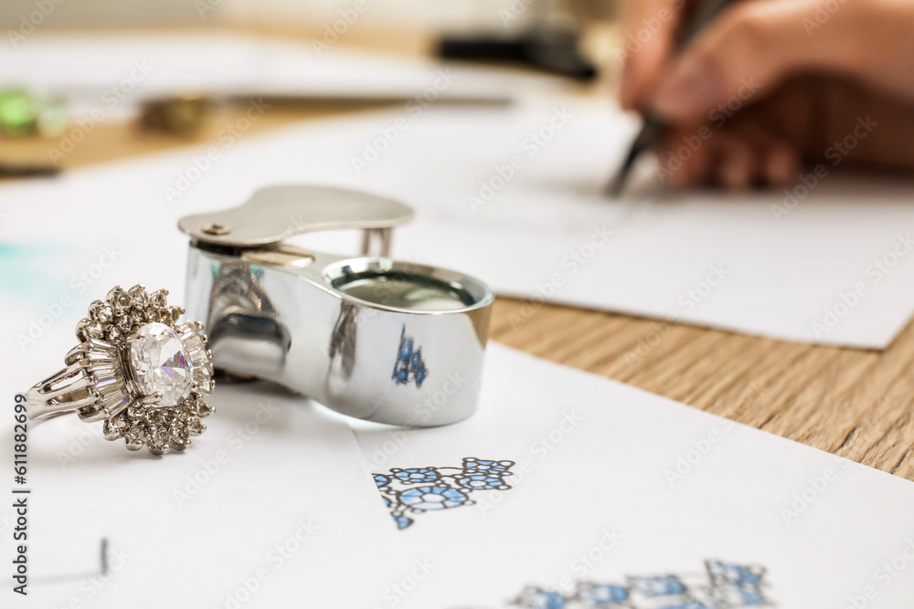 Ring with jewelers magnifier and drawings on wooden table, closeup