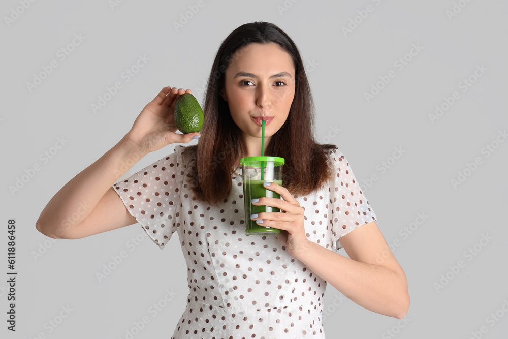 Young woman with avocado drinking vegetable juice on grey background