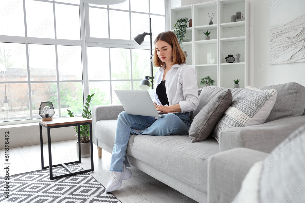 Pretty young woman sitting on grey sofa and using modern laptop in light living room