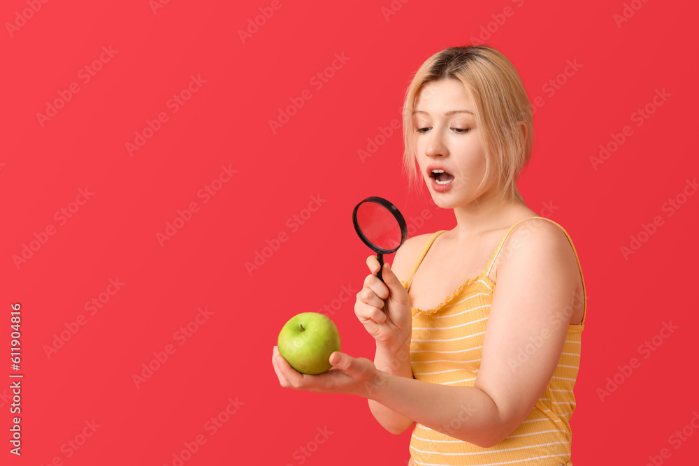 Shocked young woman with magnifier and apple on red background