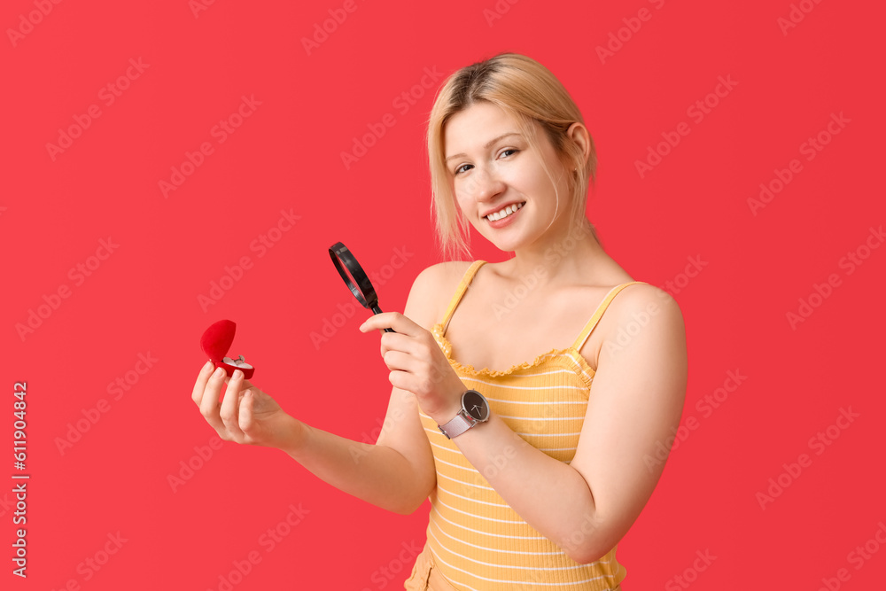 Young woman with magnifier and engagement ring on red background