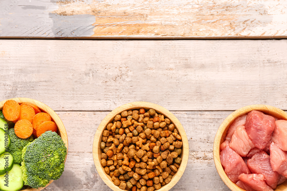 Bowls with dry pet food, raw meat and natural products on light wooden background