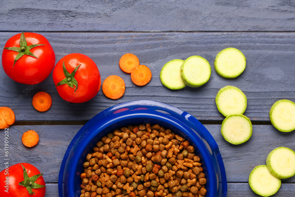 Bowl with dry pet food and natural products on color wooden background, closeup