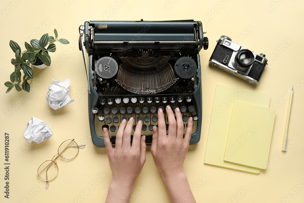 Woman typing on typewriter with notebooks, eyeglasses and camera on light yellow background