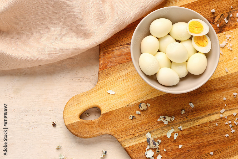 Board with bowl of boiled quail eggs and shells on white wooden background