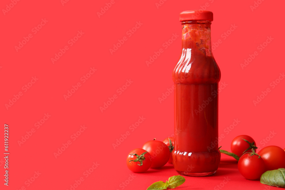 Glass bottle of ketchup and tomatoes on red background