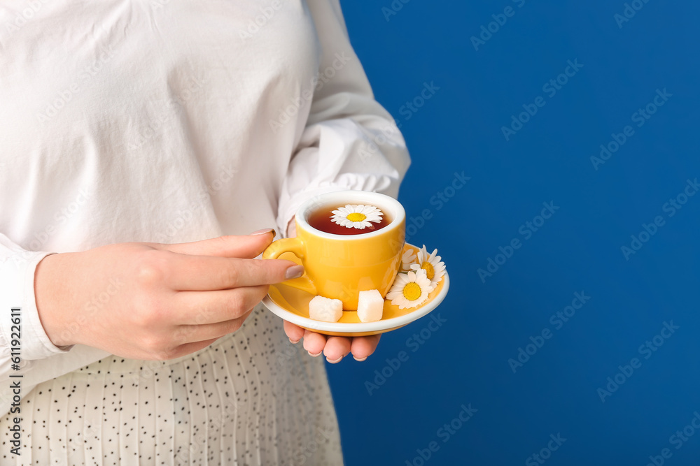 Beautiful young woman with cup of chamomile tea and flowers near blue wall