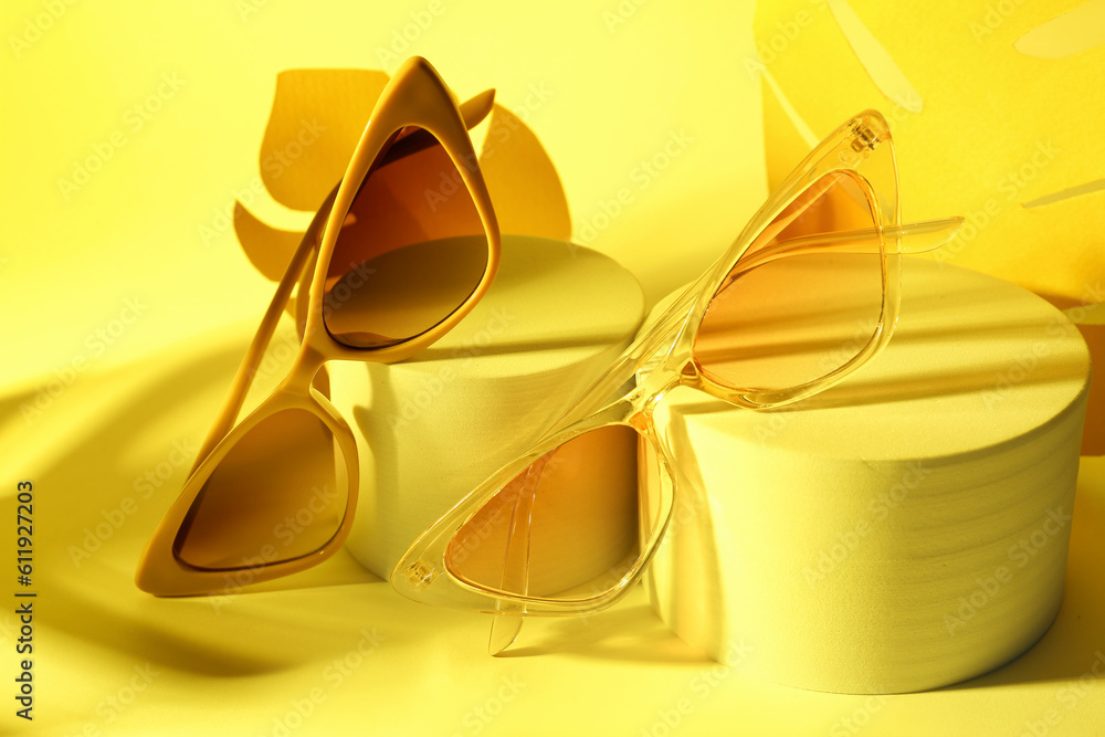 Podiums with stylish different sunglasses and paper palm leaves on yellow background