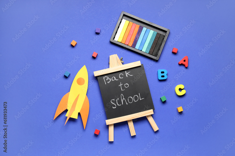 Board with text BACK TO SCHOOL, paper rocket and colorful chalks on dark blue background