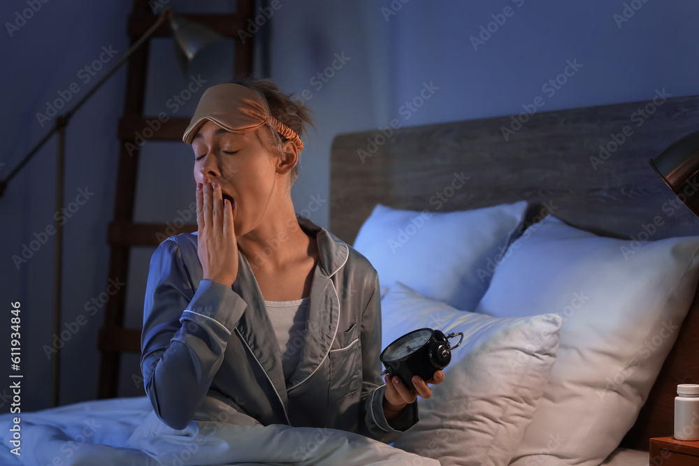 Sleepy young woman with alarm clock in bedroom at night