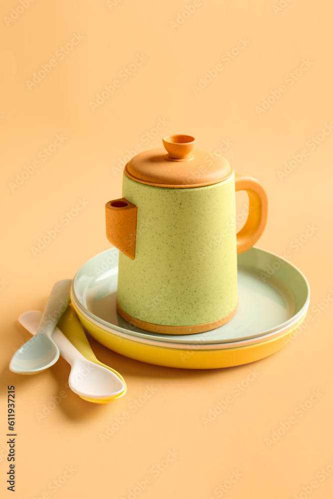 Teapot with plates and spoons for baby on pale orange background