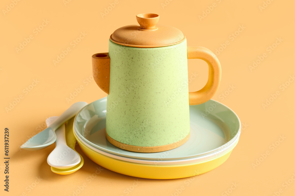 Teapot with plates and spoons for baby on pale orange background