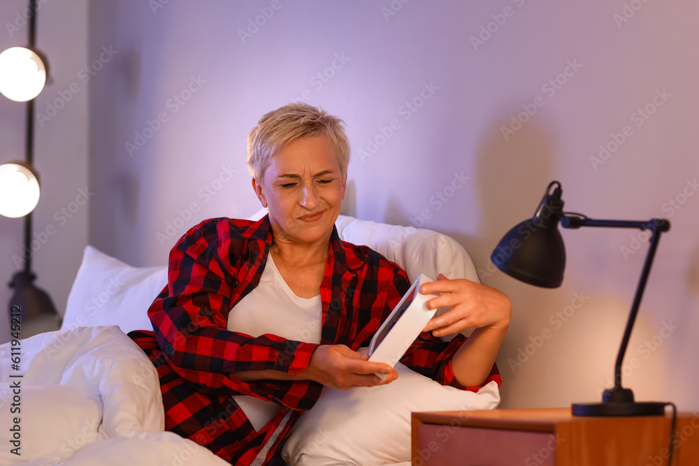 Displeased mature woman with clock in bed at night