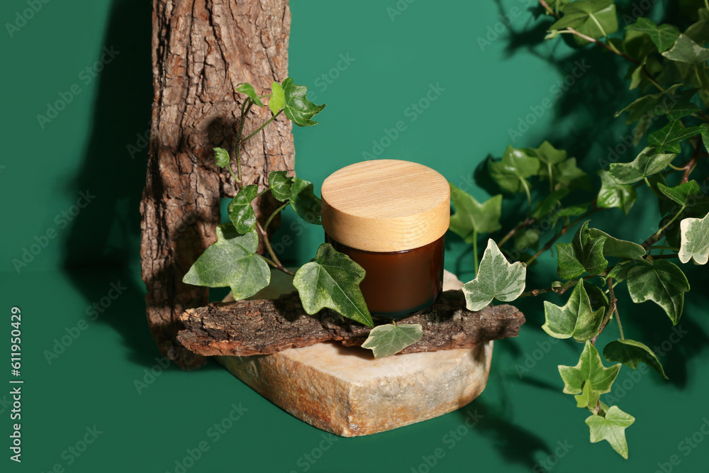 Composition with jar of cosmetic product, tree bark and ivy branch on green background