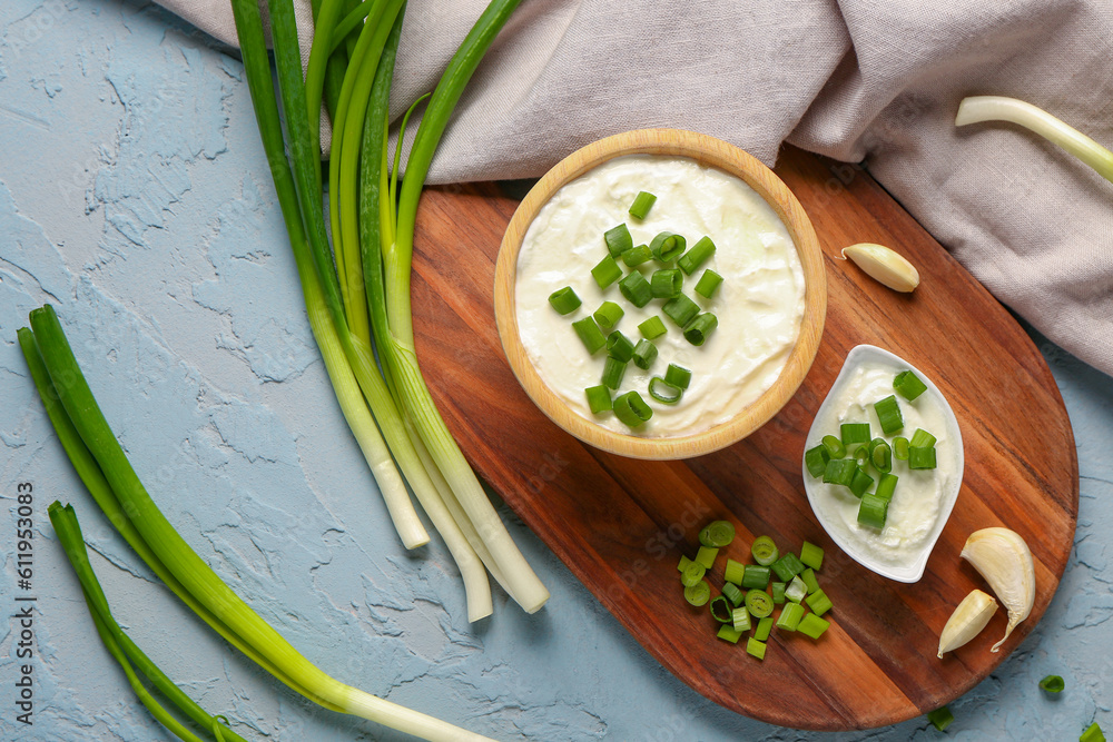 Bowl and gravy boat of tasty sour cream with sliced green onion on blue background