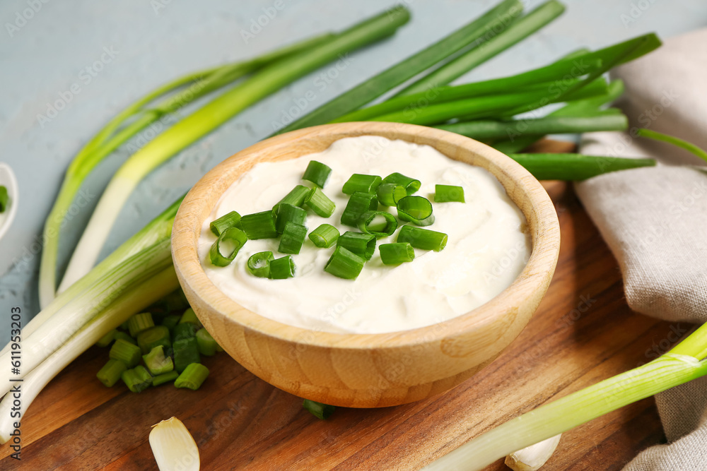 Board and bowl of tasty sour cream with sliced green onion on blue background